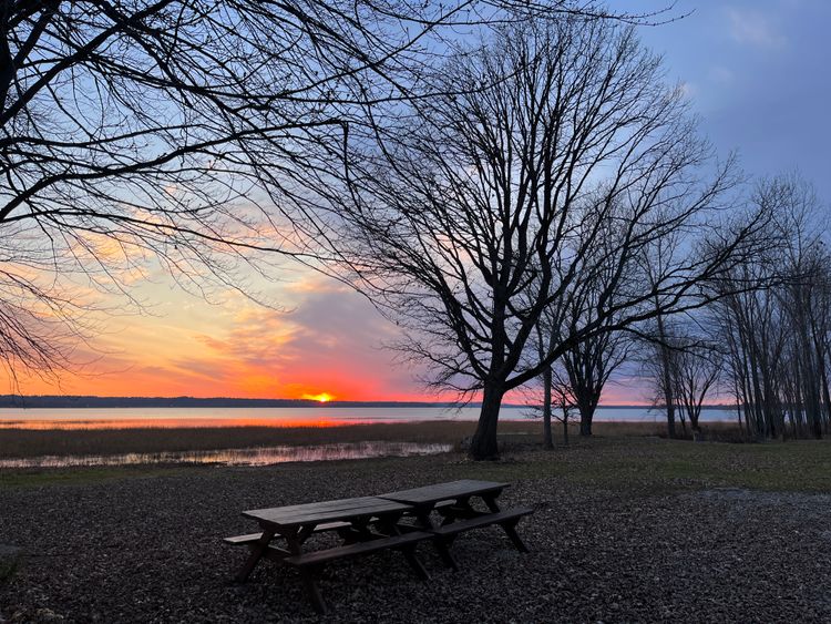 A colourful sunset over the Outaouais River framed by bare tree branches, with two empty picnic tables in foreground.